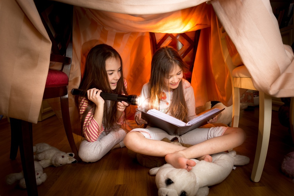 Duas meninas lendo um livro embaixo de uma cabaninha no quarto - foto: kryzhov/ShutterStock.com